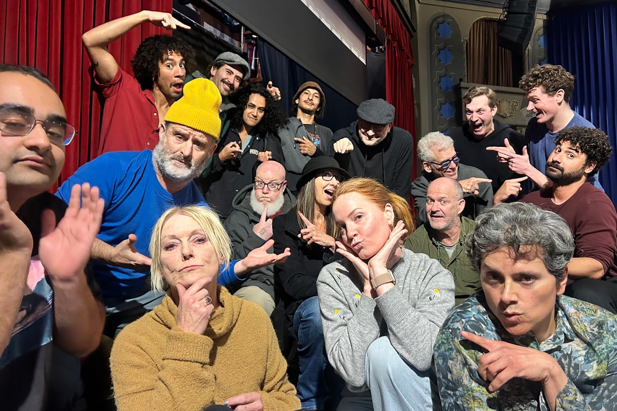 A diverse group of people poses playfully together, showcasing a variety of expressions and gestures against a backdrop of red curtains.