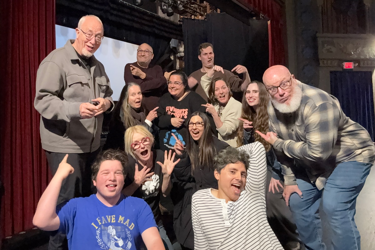 A lively group of people posing together, expressing joy and humor in a theater setting with vibrant red curtains in the background.